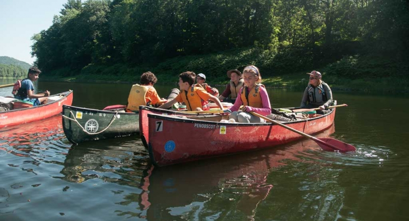 A group of people wearing lifejackets sit in canoes floating on calm water 